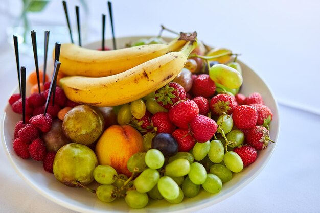 Assiette avec des fruits frais savoureux tels que des bananes, des fraises, des raisins, des pêches et des poires. Nourriture végétarienne Bol de baies mélangées sur la table