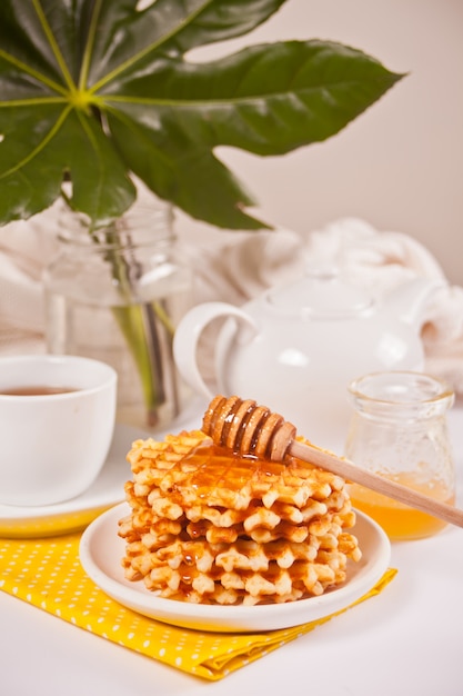 Assiette avec des gaufres savoureuses avec du miel, des baies, une tasse de thé sur la surface blanche