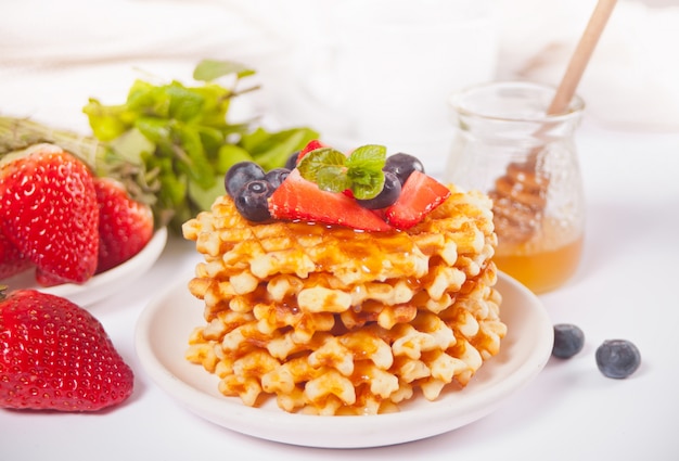 Assiette avec des gaufres savoureuses avec du miel, des baies, une tasse de thé sur la surface blanche