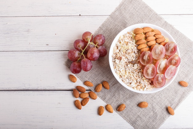 Une assiette avec muesli, amandes, raisins roses sur un fond en bois blanc.