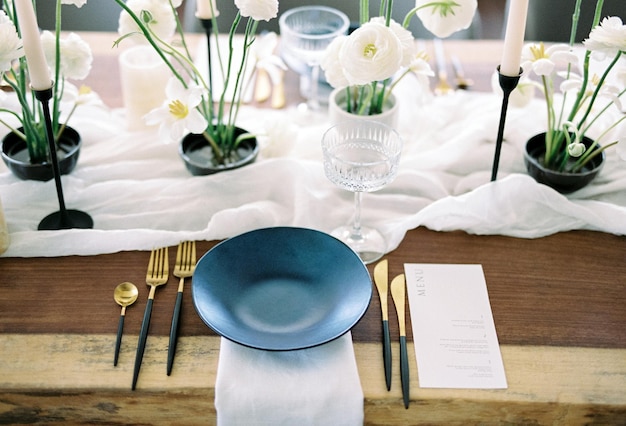 Une assiette noire se tient sur une serviette à côté de fleurs blanches sur une nappe étroite sur une table festive