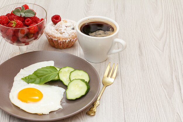 Assiette avec œuf au plat, concombre frais coupé et feuille de basilic, fourchette, tasse de café noir, muffin et bol en verre avec framboises fraîches sur fond de bois gris