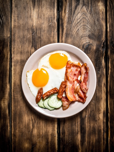 Assiette d'œufs au plat, bacon, concombre et saucisses fumées sur une table en bois