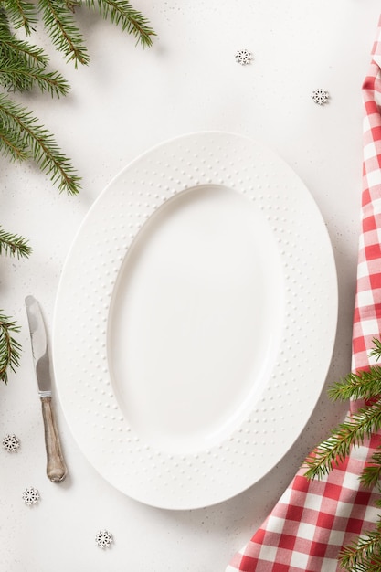 Assiette ovale vide de Noël avec des branches à feuilles persistantes de serviette rouge à carreaux sur la verticale blanche