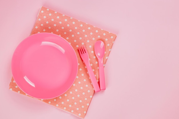 Assiette en plastique de couleur rose avec cuillère en plastique, fourchette et tissu à pois rose sur fond rose