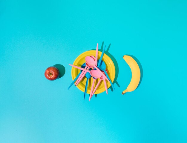 Assiette en plastique jaune avec cuillères en plastique bleu et rose et pomme et banane sur les côtés du bac bleu