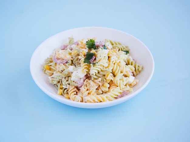 Assiette de salade de pâtes avec du maïs, du fromage et de la confiture, du persil sur le dessus, vue horizontale sur une surface bleue avec copie espace