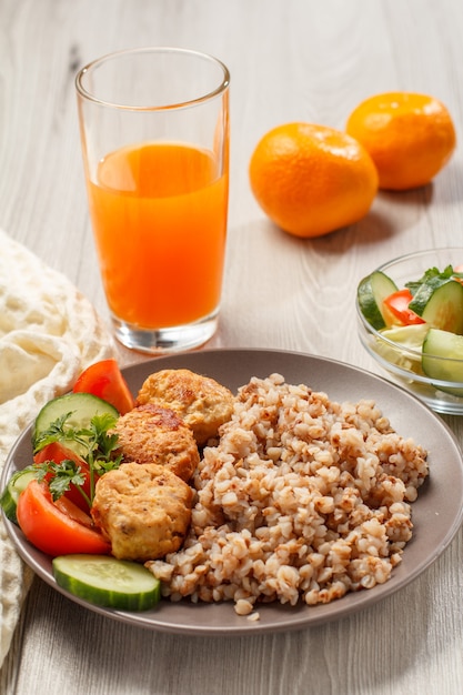 Assiette de sarrasin, escalopes de viande frites, morceaux de concombres frais, tomates et persil frais, fourchette, salade de concombres frais et tomates dans un bol en verre, verre de jus d'orange et d'oranges.