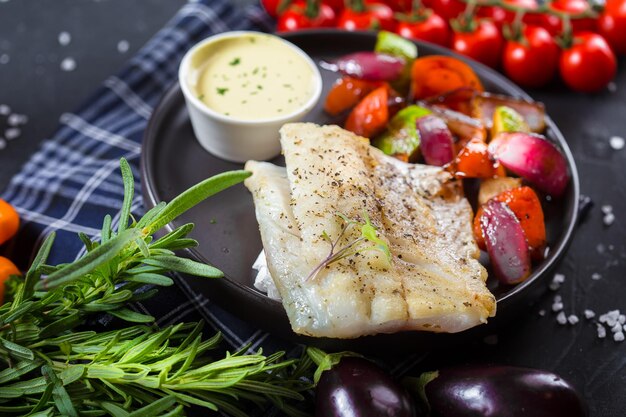 Photo sur une assiette se trouve un steak de poisson frit avec des légumes, des tomates cerises, des couverts sur un noir isolé