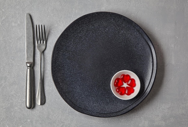 Une assiette sombre vide avec des couverts et des coeurs au caramel rouge dans un bol sur une table en béton clair Présentation romantique Vue de dessus mise à plat