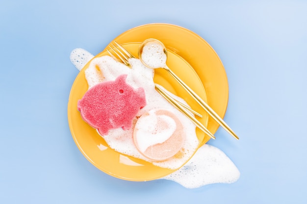 Assiettes jaunes avec cuillères, fourchettes et éponge sur fond de mousse savonneuse. Concept de lavage de vaisselle. Mise à plat, vue de dessus.