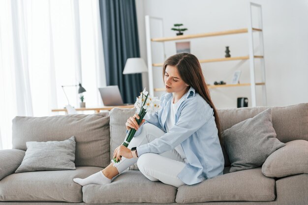 Assis sur un canapé avec des fleurs dans les mains Belle jeune femme est seule à la maison dans la chambre domestique