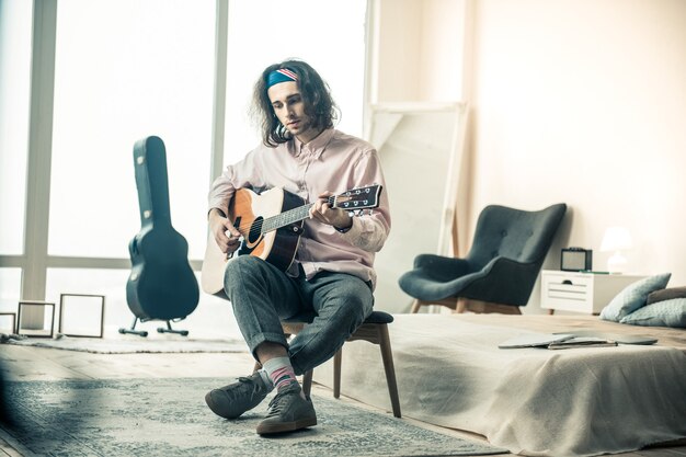 Assis dans la chambre. Beau musicien mélancolique aux cheveux ondulés jouant de la guitare acoustique tout en améliorant ses compétences