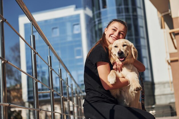 Assis ensemble Une jeune femme positive s'amuse avec son chien lorsqu'elle se promène à l'extérieur près d'un bâtiment commercial