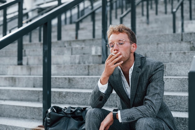 Assis et fume une cigarette Jeune homme d'affaires prospère en tenue de soirée grise est à l'extérieur de la ville