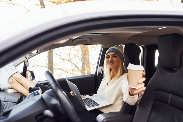 Assis avec un ordinateur portable et une tasse de boisson. La fille a un voyage d'automne en voiture. Nouvelle automobile moderne dans la forêt.