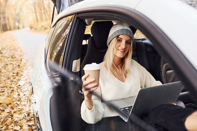 Assis avec un ordinateur portable et une tasse de boisson. La fille a un voyage d'automne en voiture. Nouvelle automobile moderne dans la forêt.