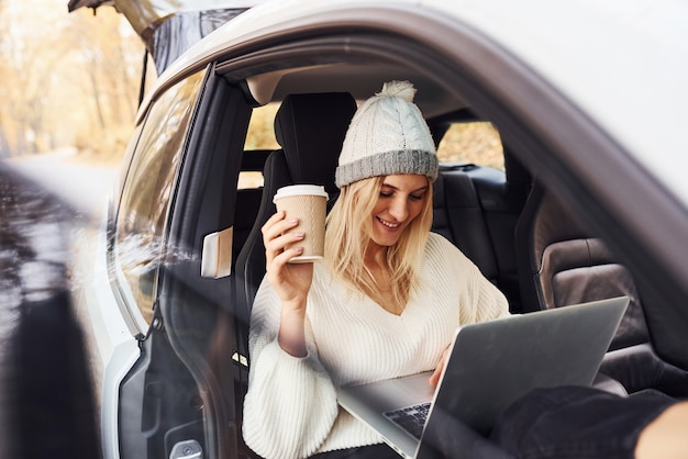 Assis avec un ordinateur portable et une tasse de boisson. La fille a un voyage d'automne en voiture. Nouvelle automobile moderne dans la forêt.