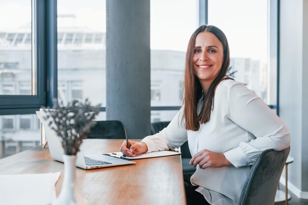 Assis près de la table Jeune femme en vêtements formels blancs est à l'intérieur dans le bureau moderne