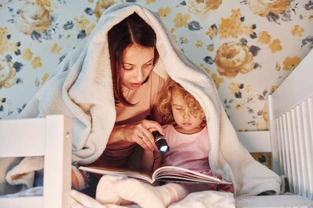 Assis sous une couverture avec une lampe de poche Jeune mère avec sa petite fille dans des vêtements décontractés ensemble à l'intérieur à la maison