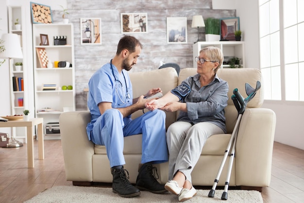 Assistance masculine faisant la surveillance de la pression artérielle pour une femme âgée dans une maison de retraite assise sur un canapé. Béquilles pour vieille femme.