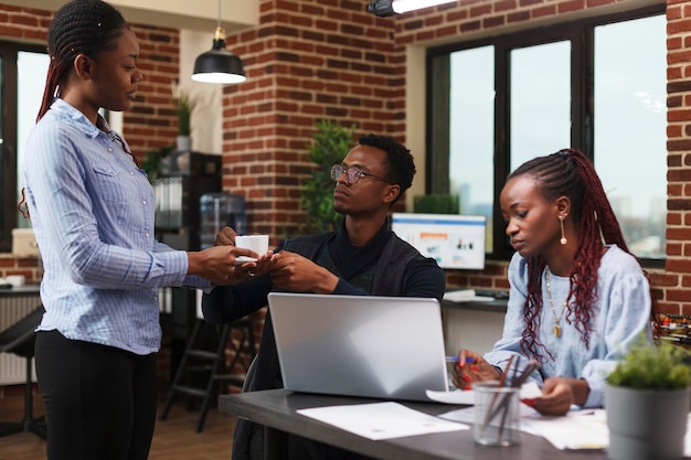 Assistant afro-américain offrant au chef de projet une tasse de café dans une salle de réunion moderne. Employés de bureau discutant des données financières et du plan de gestion des activités de recherche.