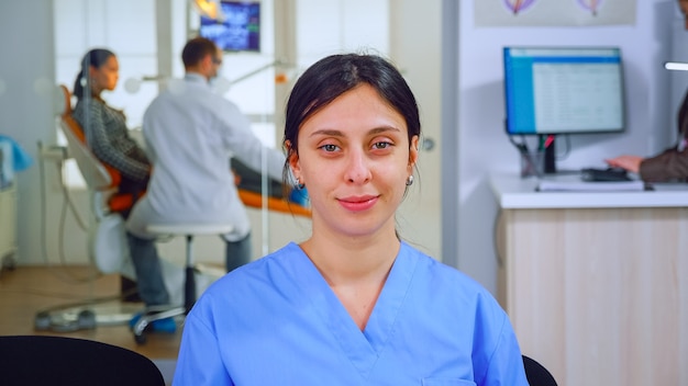 Assistant dentaire regardant la caméra pendant que le médecin examine le patient en arrière-plan. Infirmière stomatologue professionnelle souriante sur webcam assise sur une chaise dans la salle d'attente de la clinique stomatologique.