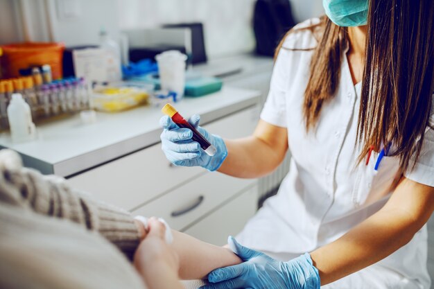 Assistant de laboratoire caucasien en uniforme, avec masque et gants en caoutchouc tenant le tube à essai avec du sang tandis que le patient est assis et tenant du coton absorbant.