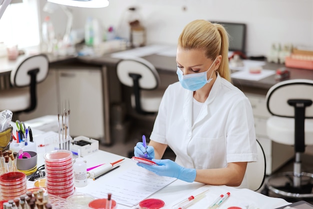 Assistant de laboratoire dédié avec masque de protection et gants sur l'écriture sur les résultats des tests de boîtes de Pétri.