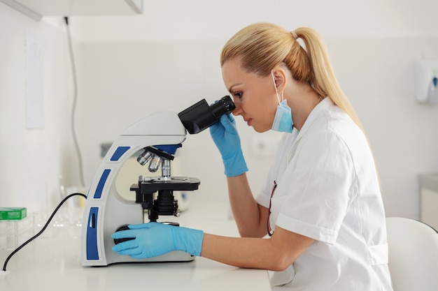 Assistant de laboratoire femme blonde assise en laboratoire et à l'aide d'un microscope pour un test sanguin.