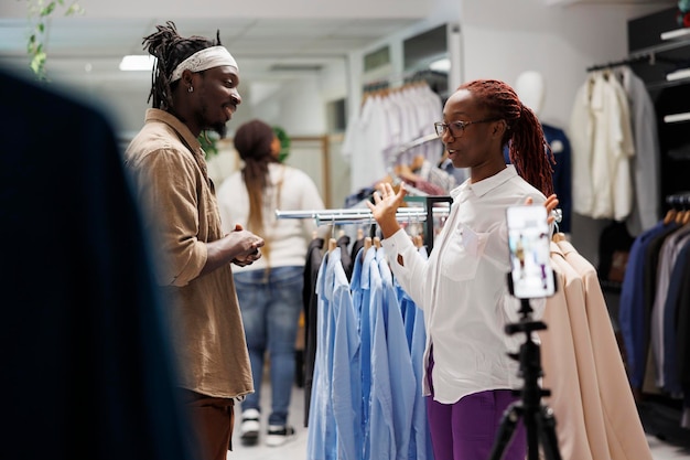 Assistant de magasin de vêtements donnant une interview à un blogueur de mode tout en enregistrant une vidéo promotionnelle sur smartphone. Influenceur afro-américain et tenue publicitaire pour les médias sociaux
