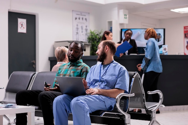 Assistant masculin parlant à un patient masculin du diagnostic, montrant les résultats de la maladie sur un ordinateur portable dans le hall de réception de l'hôpital. Spécialiste et malade discutant de médecine dans la salle d'attente.