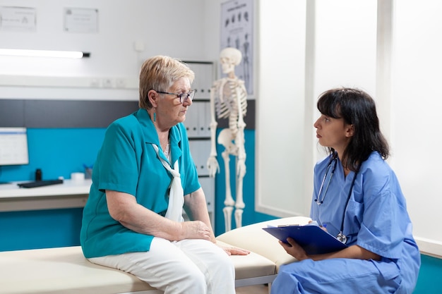 Assistant médical consultant une femme âgée blessée au bureau. Infirmière faisant un examen pour le diagnostic et la récupération orthopédiques, parlant de soulagement de la douleur et de physiothérapie