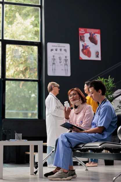 Assistant médical donnant des pilules de bouteille à un patient blessé expliquant le traitement médicamenteux lors d'un rendez-vous médical. Femme âgée portant un collier cervical ayant une consultation dans le hall de l'hôpital
