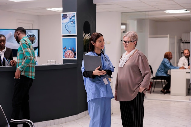 Assistant médical parlant de la maladie avec un patient âgé dans le hall d'accueil de l'hôpital, montrant un diagnostic sur un ordinateur portable pour donner un traitement de récupération. Infirmière consultant une femme âgée à la clinique médicale.