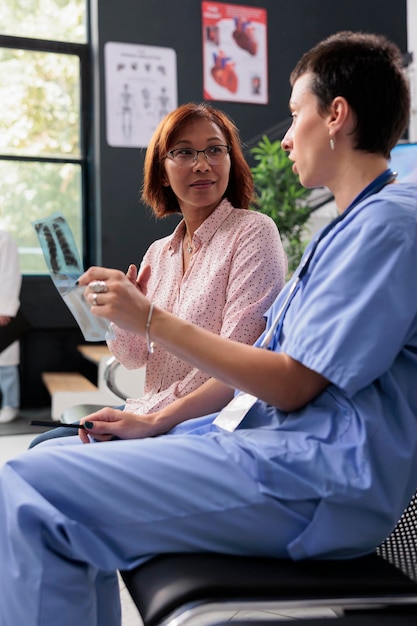 Assistant médical tenant une radiographie des poumons expliquant l'expertise au patient âgé dans la zone d'attente de l'hôpital. Infirmière discutant d'un traitement de santé lors d'une consultation clinique