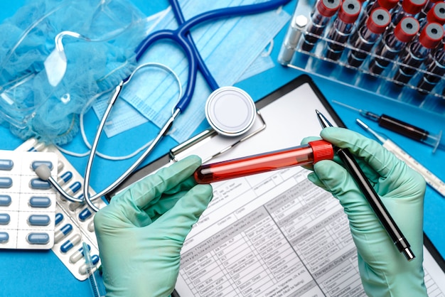Assistant technicien de laboratoire ou médecin portant des gants en caoutchouc ou en latex tenant le tube à essai sanguin sur le presse-papiers avec un formulaire vierge.