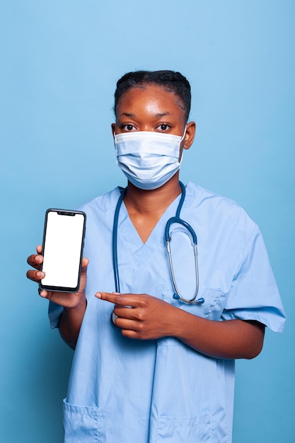 Assistant thérapeute afro-américain portant un masque de protection pour prévenir l'infection par le coronavirus tenant un smartphone avec écran blanc travaillant en studio avec fond bleu. Notion de médecine