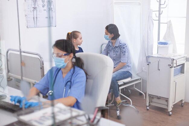 Assistant thérapeute écrivant un traitement sur un ordinateur portable pendant la consultation d'un enfant au bureau de l'hôpital Spécialiste des maladies portant un masque de protection contre le coronavirus pendant la pandémie mondiale