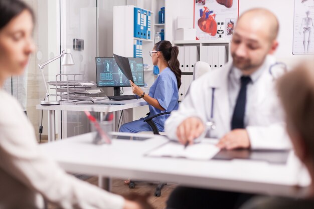 Assistant En Uniforme Bleu Regardant L'image Radiographique Dans Le Bureau De L'hôpital. Médecin Portant Une Blouse Blanche Lors De La Consultation D'une Femme âgée Handicapée En Fauteuil Roulant Expliquant Le Diagnostic à Sa Fille.