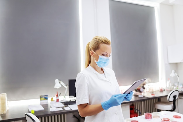 Assistante de laboratoire innovante et expérimentée en uniforme stérile blanc avec masque chirurgical et gants en caoutchouc debout dans le laboratoire et utilisant une tablette pour entrer les résultats des tests.