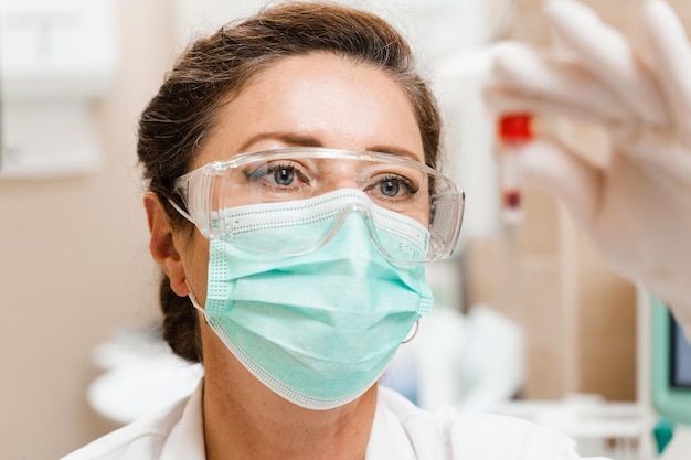 Une assistante de laboratoire tient un tube à essai avec du sang dans ses mains et le regarde Test sanguin en laboratoire Scientifique médical portant des lunettes et un masque