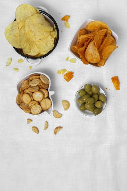 Assortiment de collations à la maison avec croustilles, bière, craquelins, olives vertes et noires sur table avec nappe en lin blanc