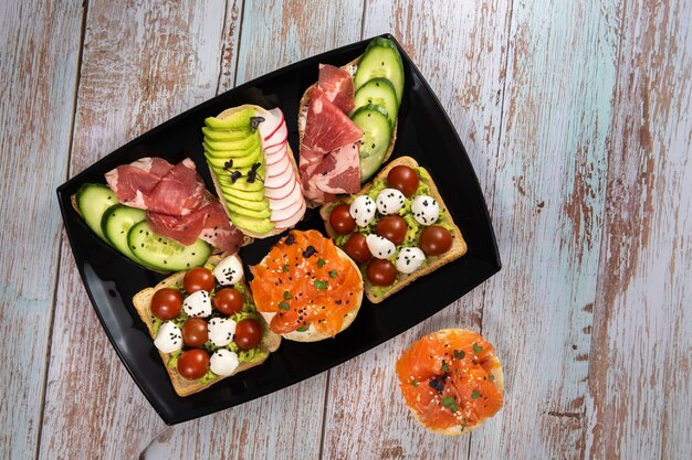 Assortiment de sandwichs avec du poisson, du fromage, de la viande et des légumes sur une plaque noire.
