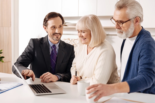 Assurer dans un choix rentable. Agent immobilier intelligent sincère optimiste rencontre avec un vieux couple de clients tout en présentant le plan de la maison et en utilisant un ordinateur portable