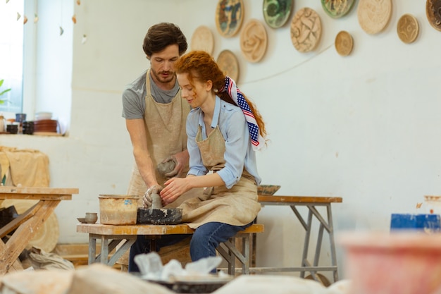 Atelier équipé. Agréable fille aux cheveux longs en tablier cloutant les aspects du travail avec un tour de potier professionnel