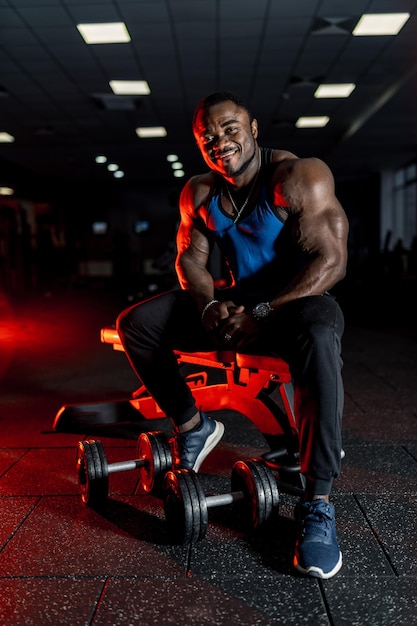 Un Athlète Africain Très Musclé Est Assis Sur Un Banc Dans Une Salle De Sport Moderne. Le Sport Comme Mode De Vie. Lumière Sombre. Fermer.