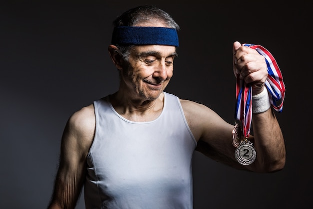 Athlète âgé portant une chemise sans manches blanche, avec des marques de soleil sur ses bras, avec trois médailles dans une de ses mains, sur fond sombre. Concept de sport et de victoire.