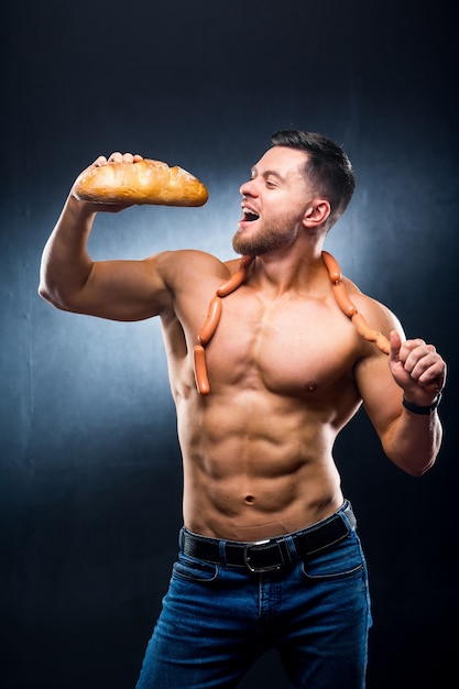 L'athlète au torse nu mange du pain frais. Saucisses sur un cou. Photo recadrée. Concept de mode de vie. Photographie d'atelier. Sportif affamé.