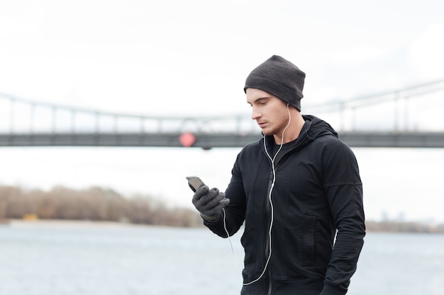 Athlète beau jeune homme en chapeau et gants écoutant de la musique avec un téléphone portable à l'extérieur